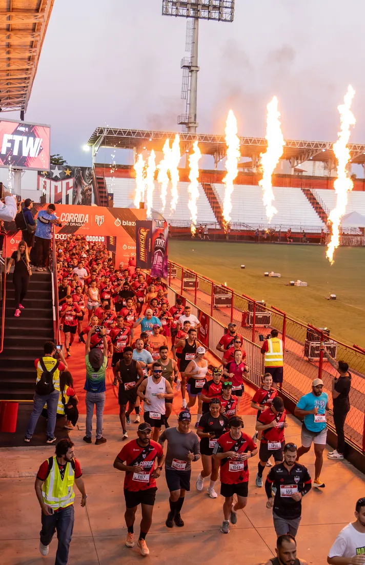Pelotão de corredores descendo para a pista do estádio com labaredas de fogo